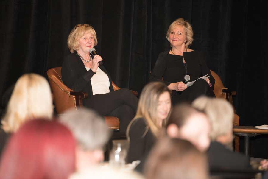 Woman with a mic talking with another woman sitting besides here